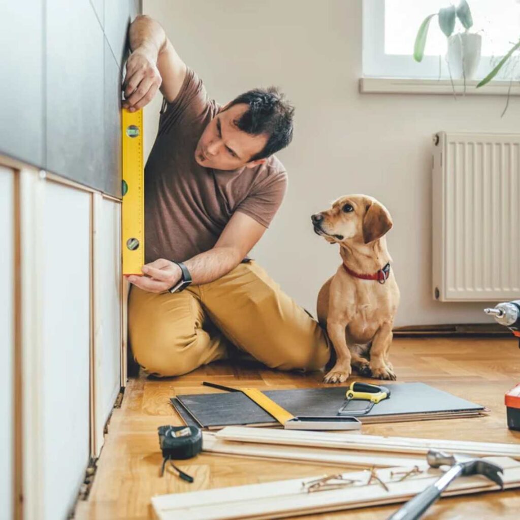 A man drilling holes measuring his wall, sat down with a dog next to him.